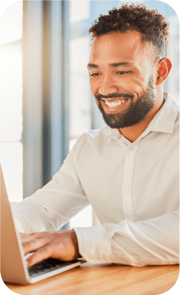 Homem de camisa branca, sorrindo enquanto mexe no computador.