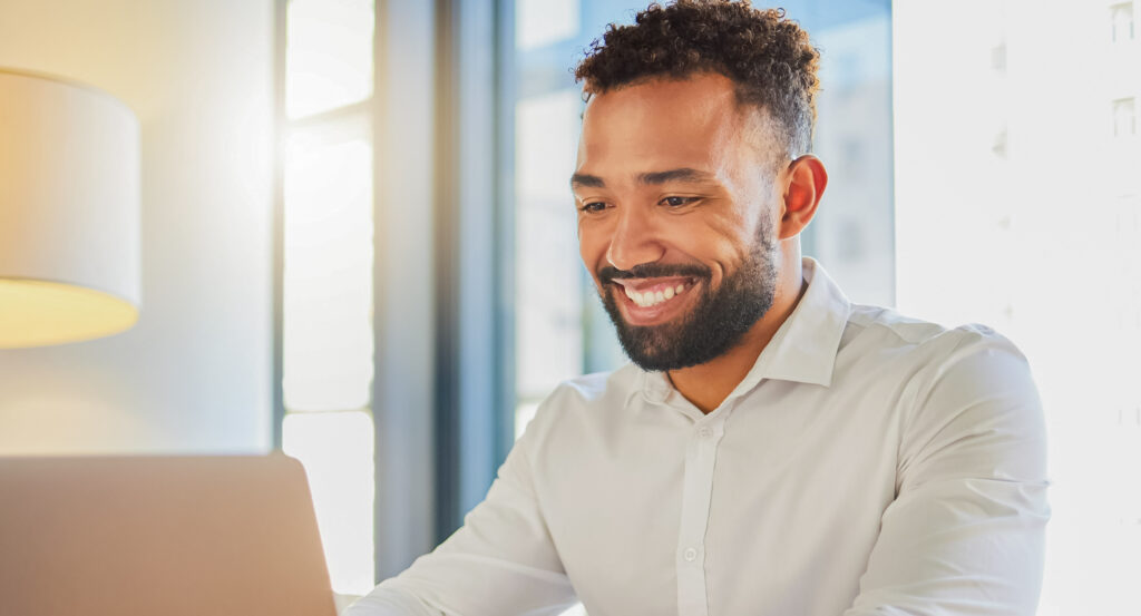 Homem de camisa branca, sorrindo enquanto mexe no computador.