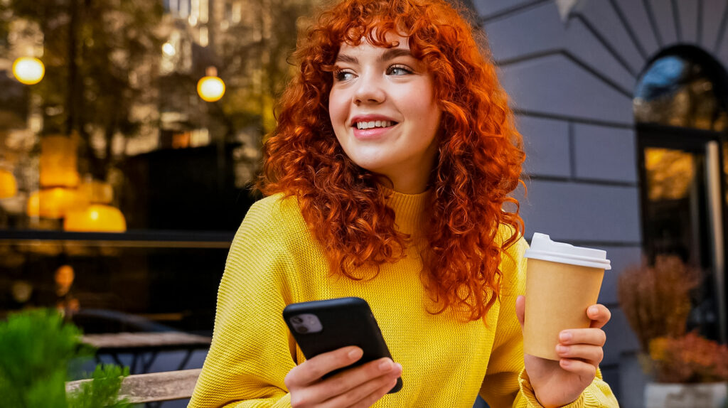 Mulher sorrindo tomando cafe, enquanto segura o celular.